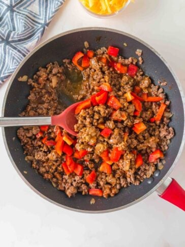 Ground beef and chopped red bell peppers cooking in a skillet with a red silicone spatula, with a small bowl of shredded cheese nearby.