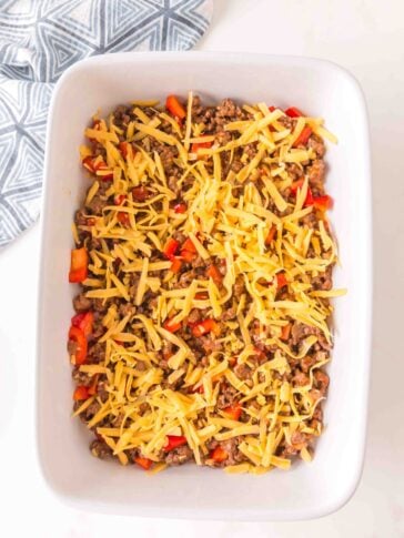 A white baking dish filled with a mixture of cooked ground meat, diced red bell peppers, and shredded cheddar cheese.