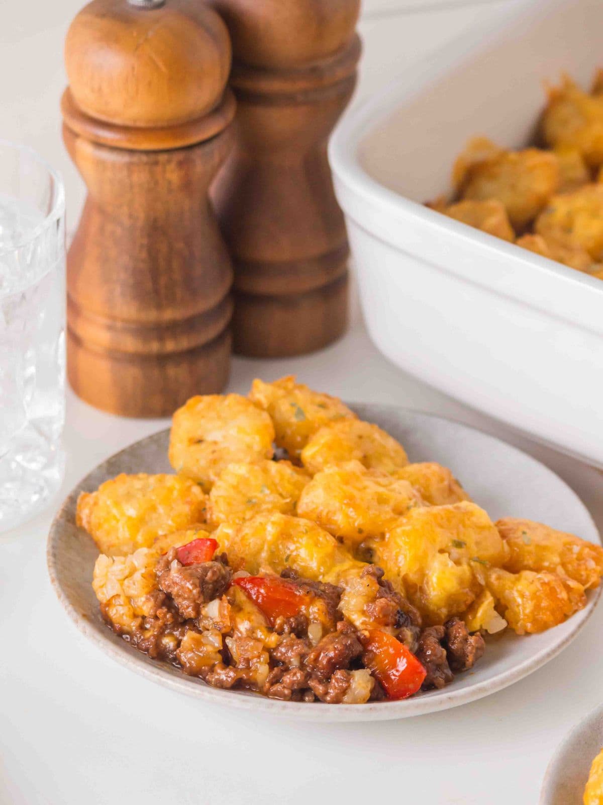 A serving of tater tot casserole with ground meat and red peppers on a small plate, with wooden salt and pepper mills in the background.