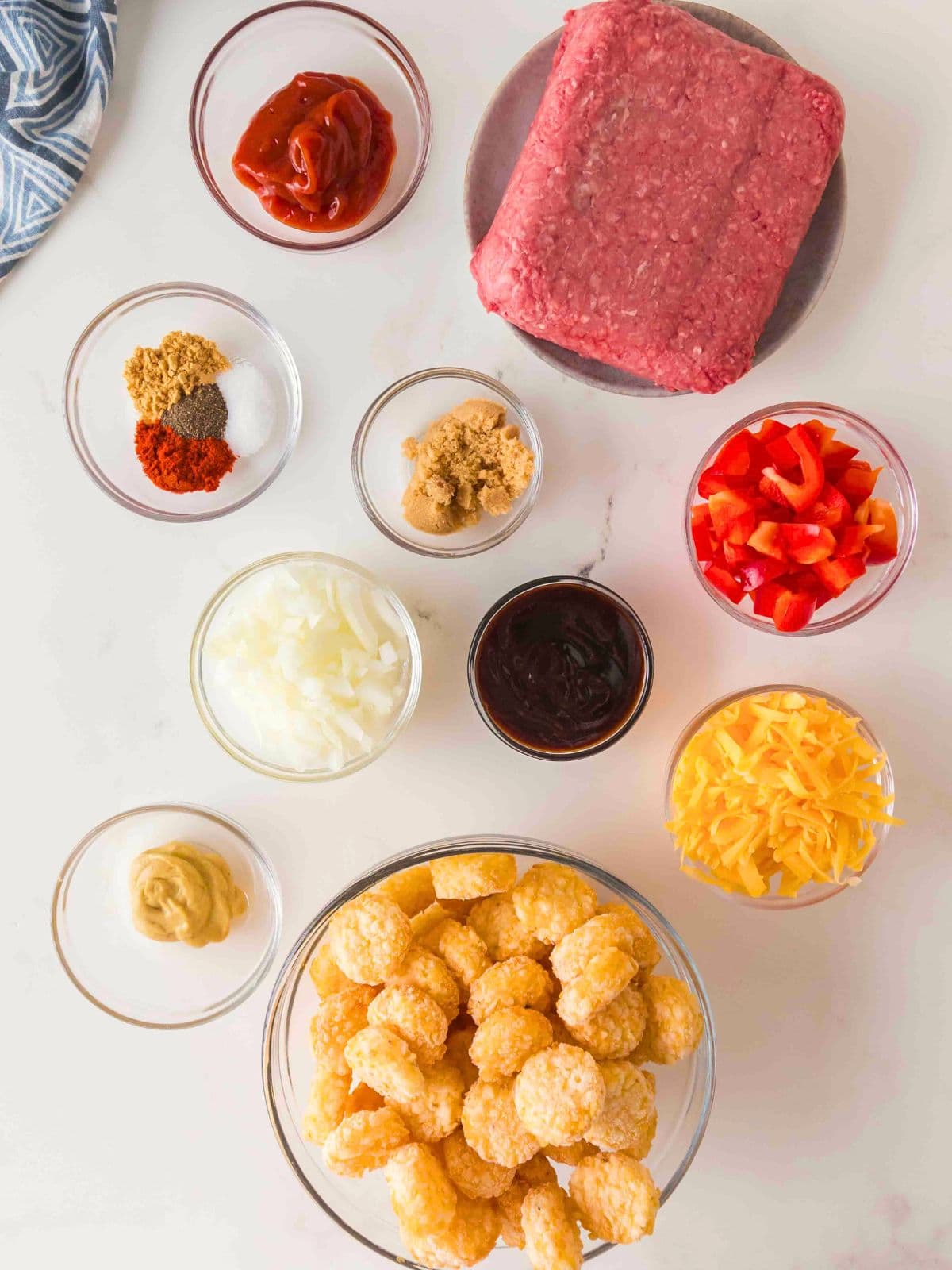 Ingredients for a recipe arranged on a white surface, including a block of ground beef, tater tots, shredded cheddar, chopped red peppers, diced onions, brown sugar, ketchup, barbecue sauce, mustard, and a small bowl of spices.