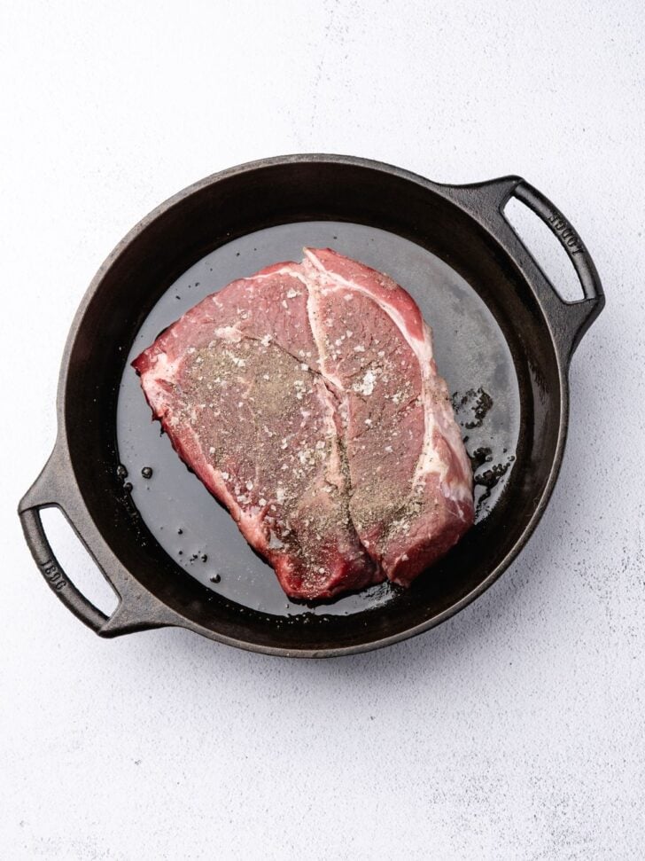 A raw steak seasoned with salt and pepper in a black cast iron skillet.