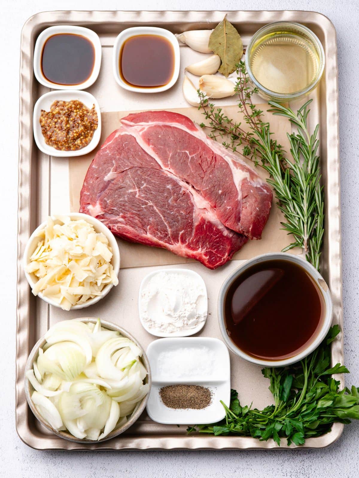 A raw beef roast on a baking sheet surrounded by bowls of sliced onions, shredded cheese, sauces, seasonings, flour, oil, garlic cloves, and fresh herbs.
