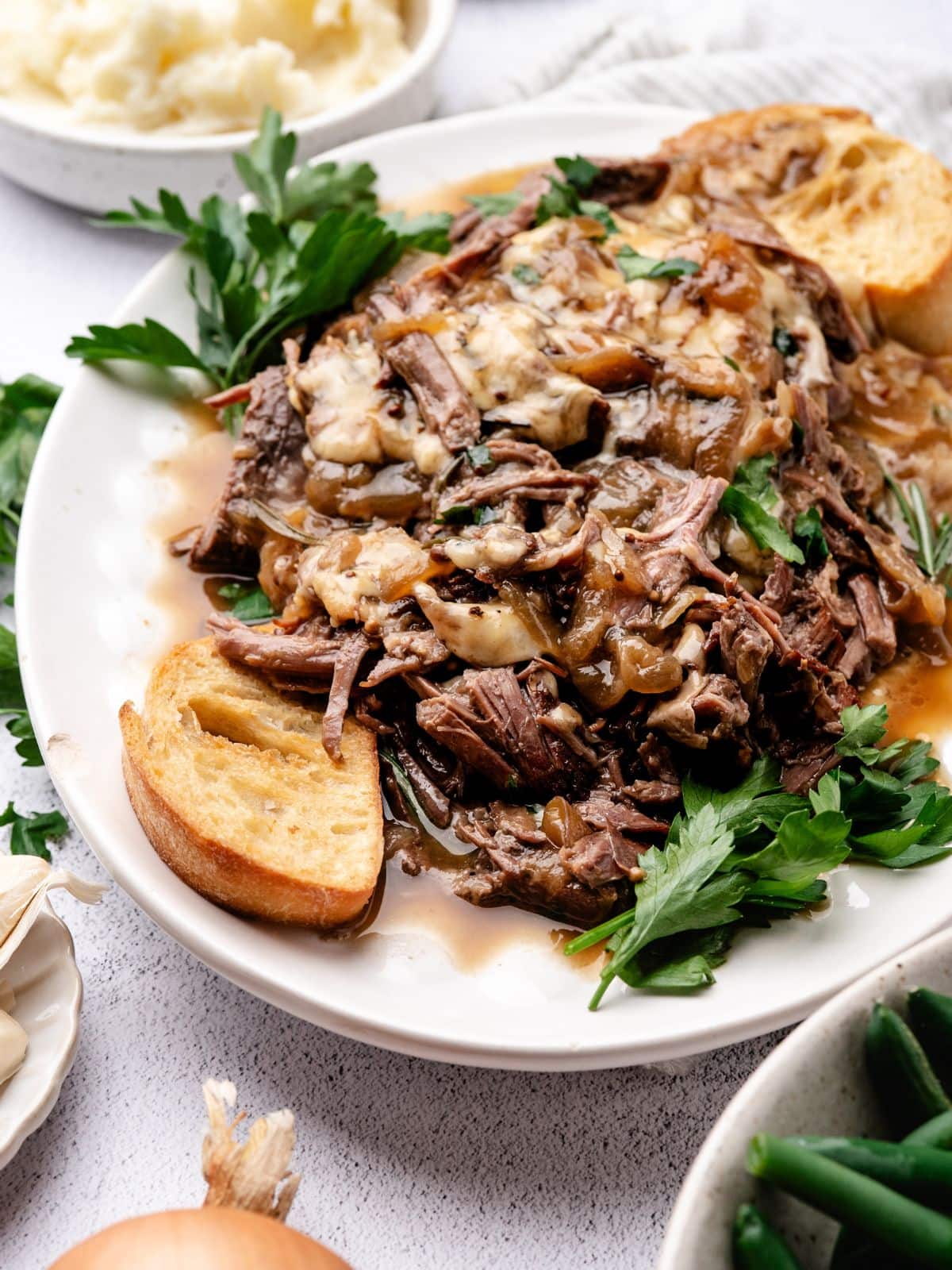 Shredded beef topped with melted cheese and caramelized onions on a plate with toasted bread and fresh parsley.
