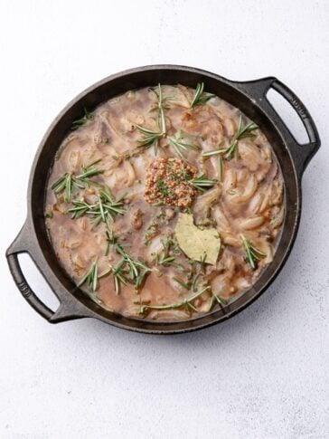 Cast iron skillet containing onion gravy garnished with rosemary sprigs, a bay leaf, and a dollop of coarse mustard.