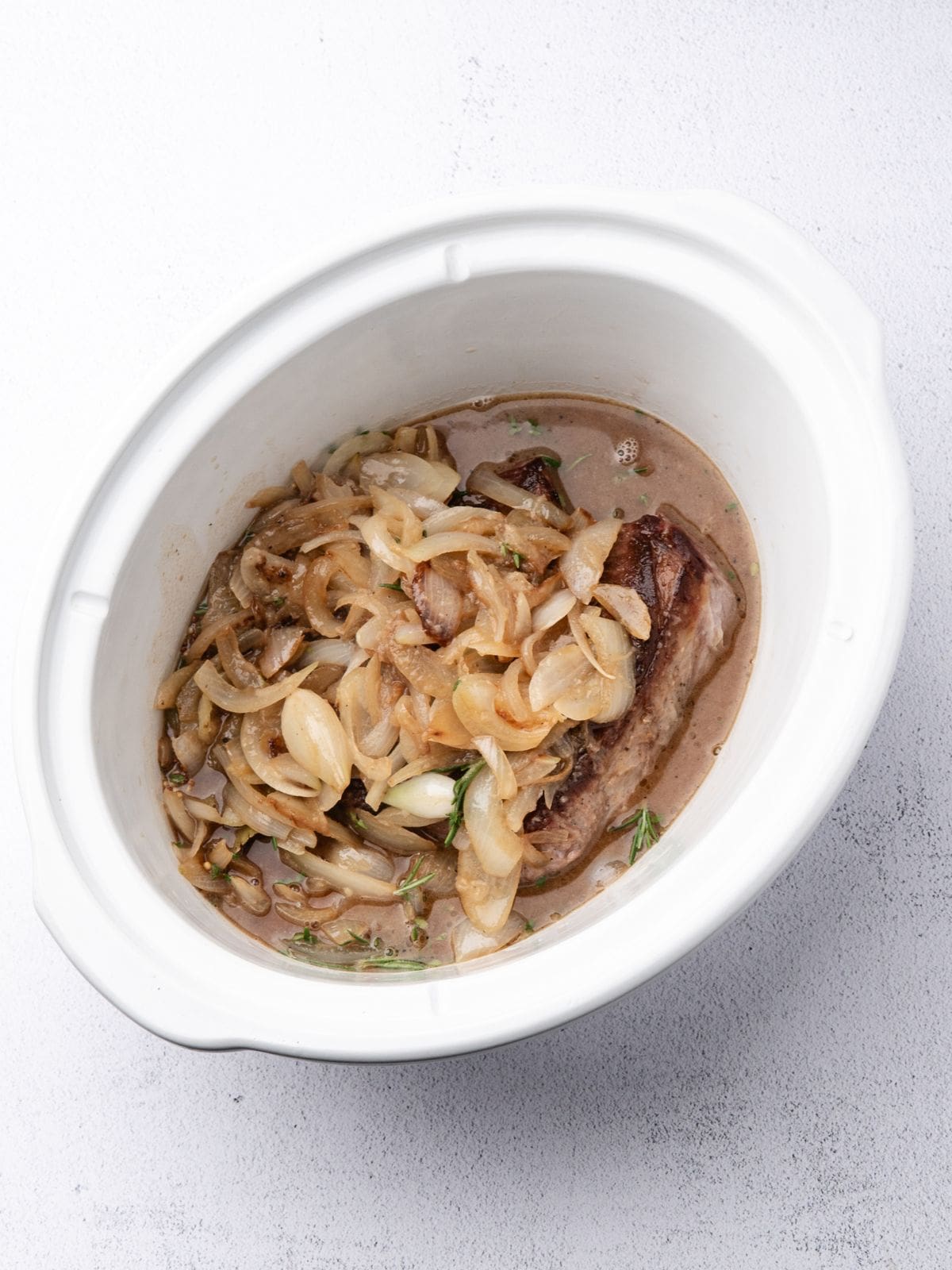 A slow cooker containing a seared piece of meat topped with sautéed onions and herbs in a brown sauce.