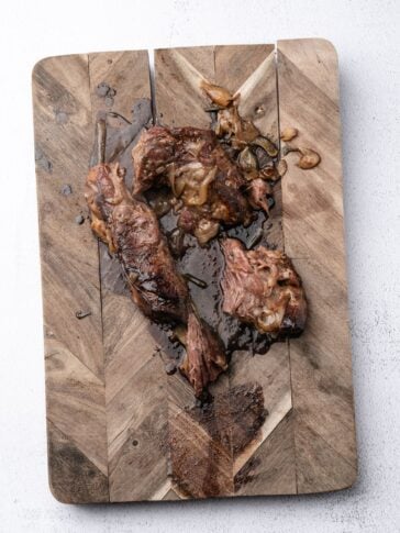 Pieces of cooked, braised meat with caramelized onions on a wooden cutting board.