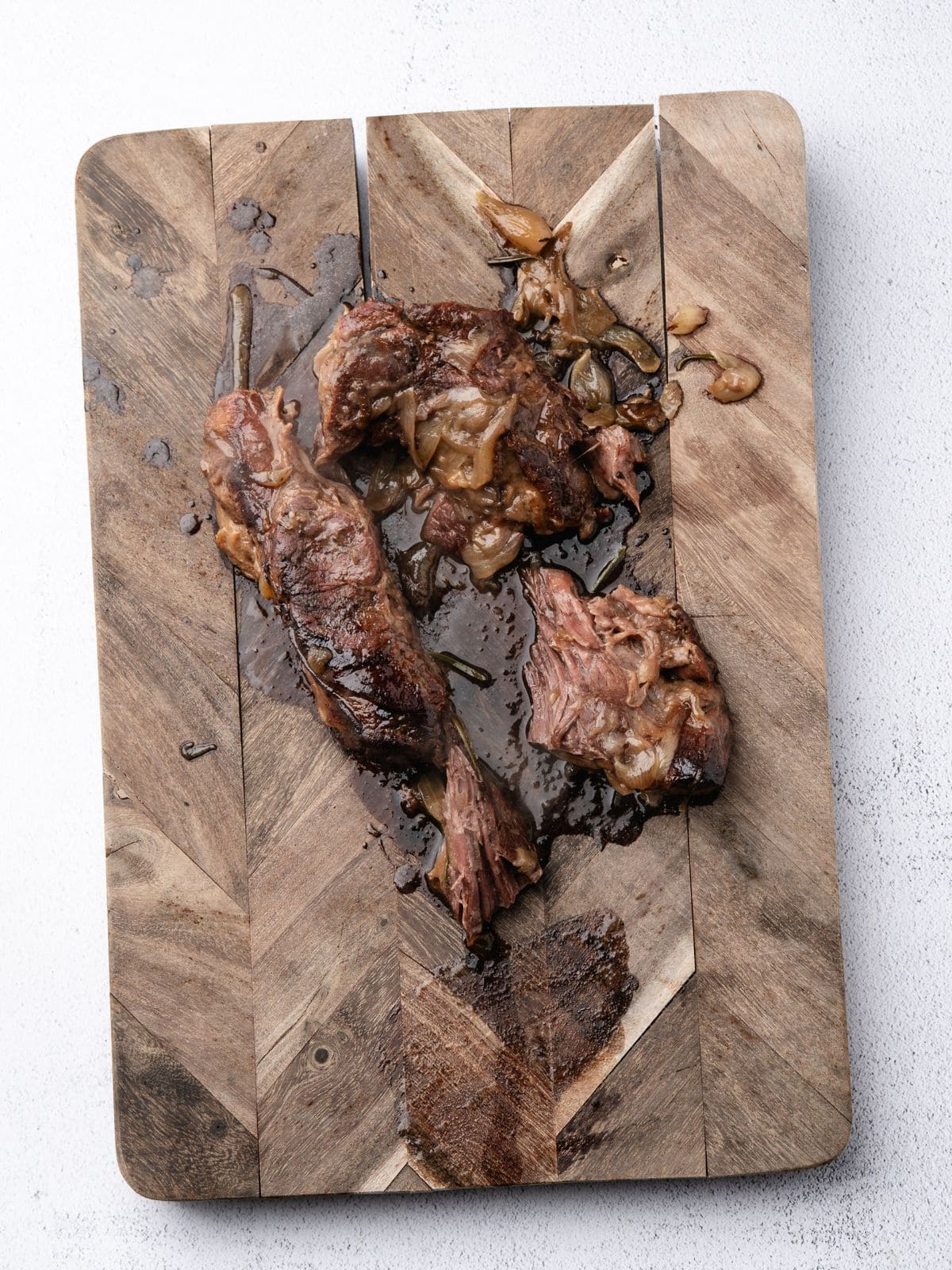 Pieces of cooked, braised meat with caramelized onions on a wooden cutting board.