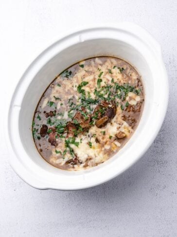 A white slow cooker filled with shredded beef in brown gravy, topped with melted cheese and fresh herbs.