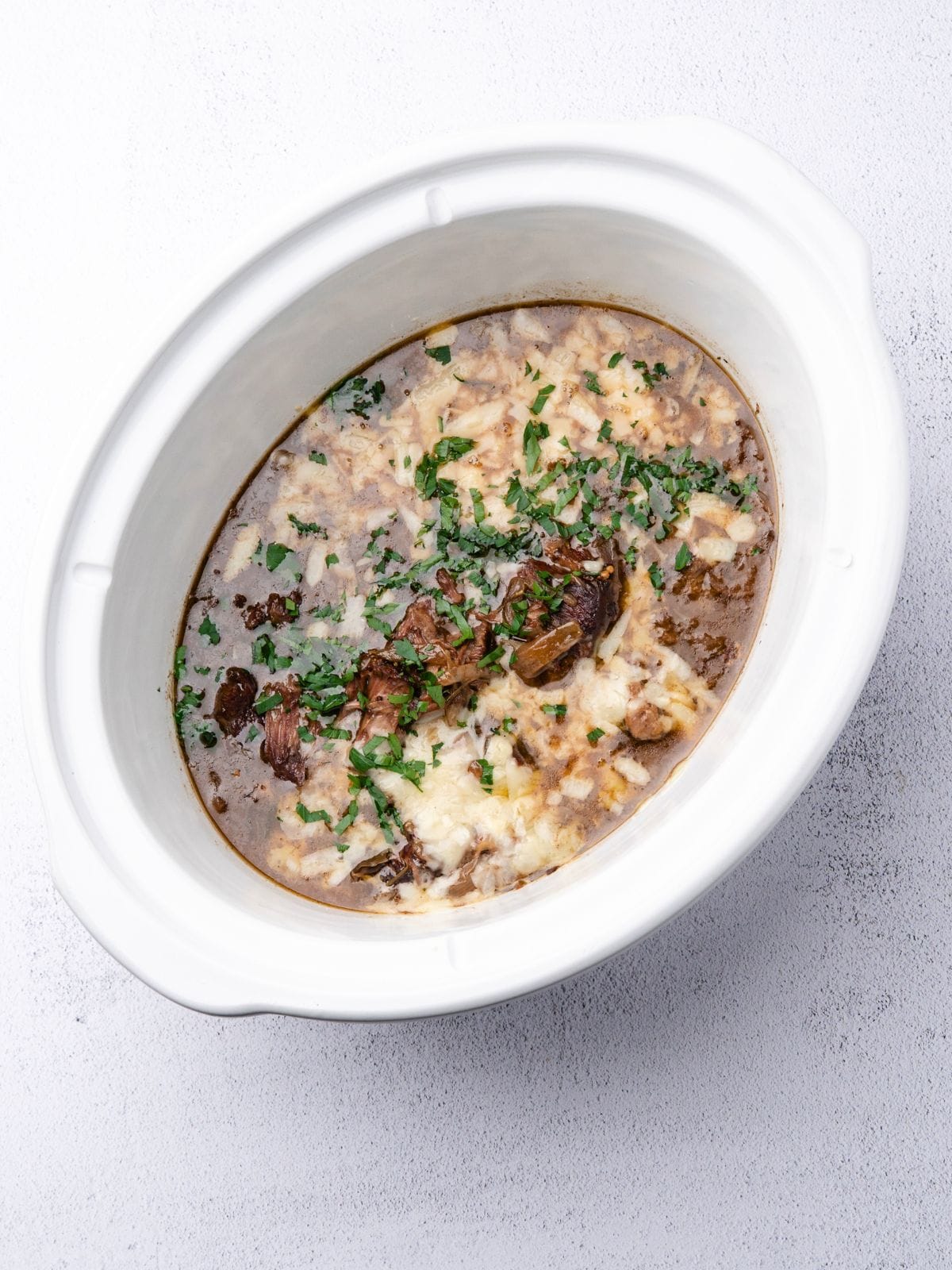 A white slow cooker filled with shredded beef in brown gravy, topped with melted cheese and fresh herbs.