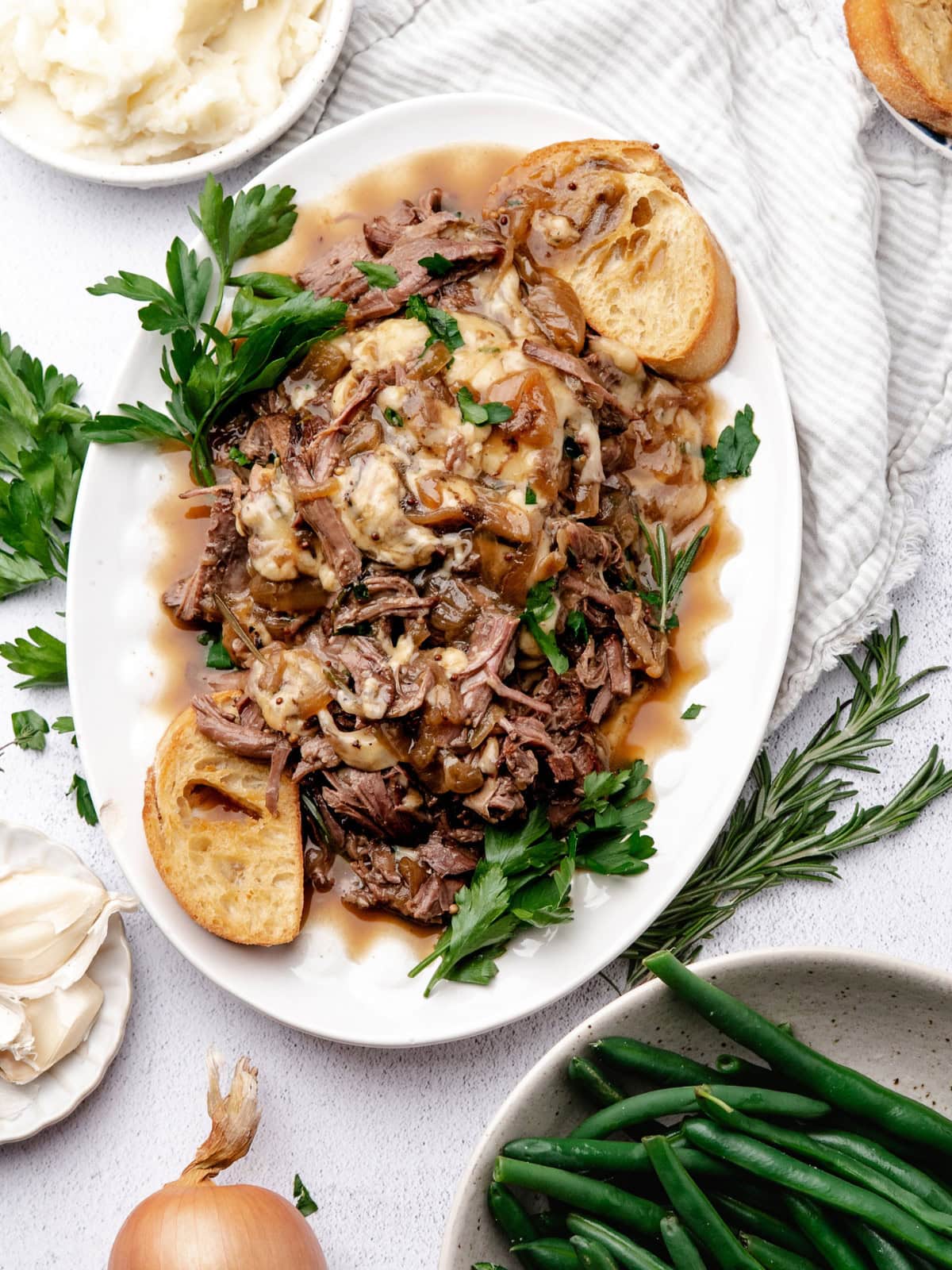 A platter of shredded beef topped with melted cheese and onions, served with toasted bread slices, fresh herbs, mashed potatoes, and green beans.