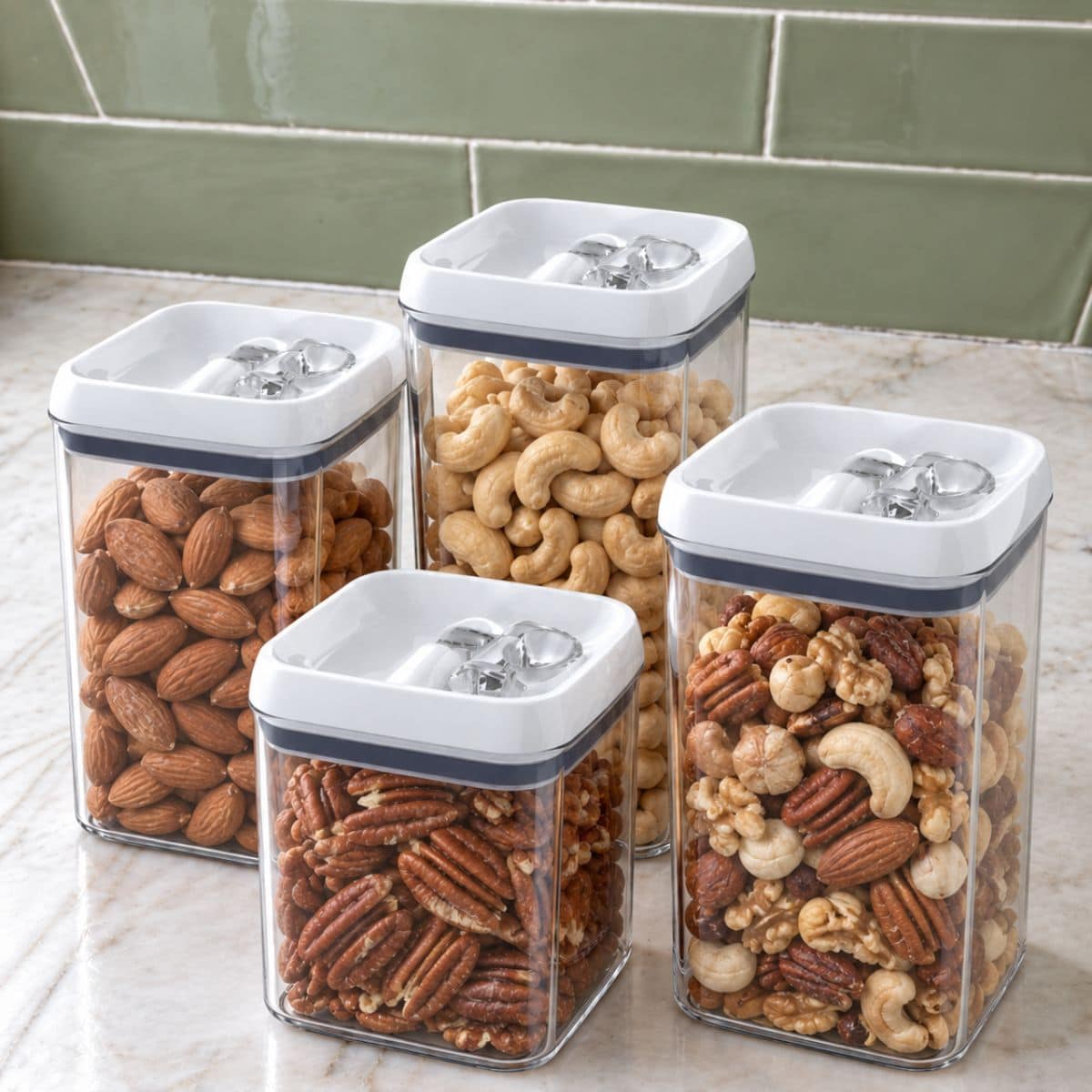 Four clear, square containers with white lids are filled with different nuts—almonds, cashews, pecans, and a nut mix. They are arranged on a marble countertop against a green tiled backsplash.