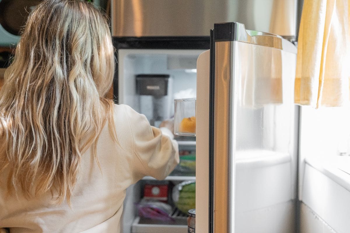 A woman with long blonde hair stands in front of an open refrigerator, reaching inside. The kitchen is well-lit by natural light from a nearby window with a yellow curtain.